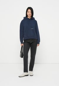 Mørkeblå hettegenser med frontlomme, snor i hetten og logodetaljer. Kombinert med svarte jeans og lyse slip-on sko.