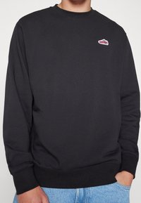 Sudadera negra con cuello redondo, puños y dobladillo acanalados. Presenta un pequeño logo multicolor en el pecho. El material parece suave y con textura.