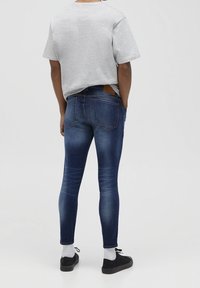 Vaqueros de denim ajustados en azul medio, con textura desgastada, cinco bolsillos y una etiqueta de cuero marrón en la parte trasera de la cintura. Combinados con una camiseta gris.