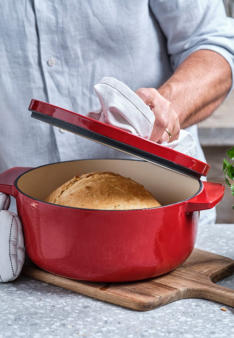  roter gusseiserner Dutch Oven auf einem Holzschneidebrett, mit einem Lid, das angehoben wird, um ein rundes Brot im Inneren zu enthüllen.
