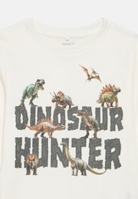 Κρεμ μπλουζάκι με γραφικό δεινόσαυρου που απεικονίζει πολλαπλά είδη και το κείμενο "ΚΥΝΗΓΟΣ ΔΕΙΝΟΣΑΥΡΩΝ" με έντονα, υφασμένα γράμματα.