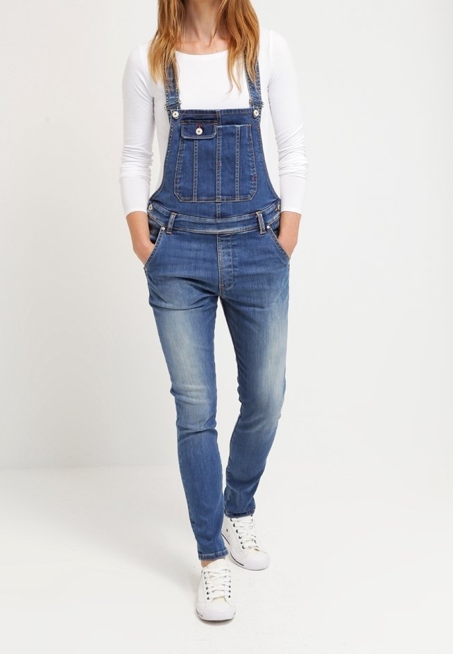 Femme portant une salopette en denim bleu avec une chemise blanche à manches longues et des baskets blanches, debout les mains dans les poches devant un fond uni.