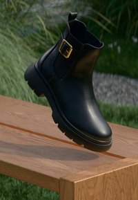 Black leather ankle boot with a shiny finish, gold buckle detail, and rugged sole. Elevated position against a wooden surface.