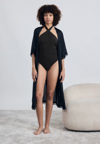 Melna caurspīdīga kimono ar mežģīņu apdari, uzvilkta virs tumši brūnas haltera stila viens gabala peldkostīma. Modelis stāv basām kājām uz gaiši teksturētas virsmas.