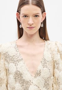 Κρεμ φόρεμα με χάντρες και υφή με λουλούδια, με βαθύ V-λαιμόκοψη. Το μοντέλο φοράει ασημένια σκουλαρίκια σε σχήμα δάκρυ, με ίσια μαλλιά μήκους μέχρι τους ώμους.