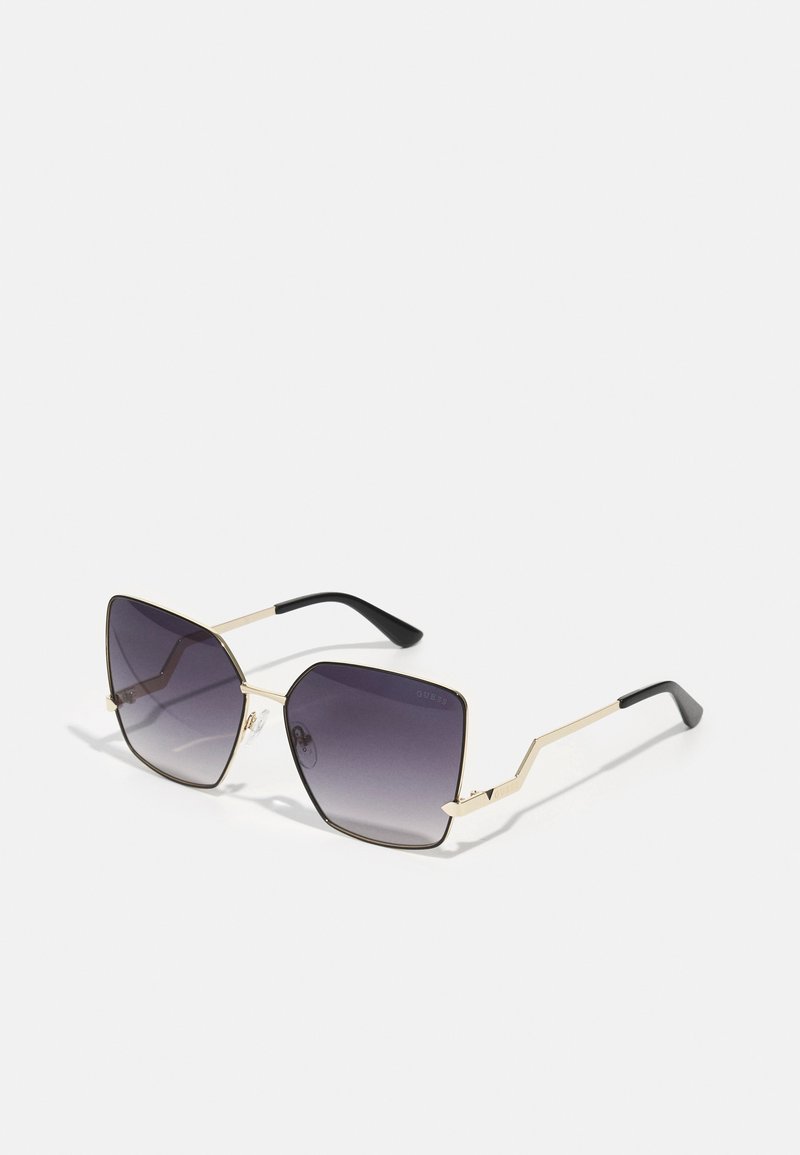 Óculos de sol oversized em preto e dourado com lentes graduadas em roxo, armação de metal angular e pontas de haste texturizadas em preto.