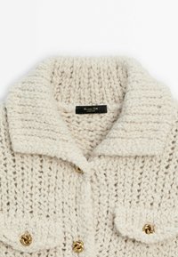 Cardigan en grosse maille crème avec large col, motif texturé et boutons nœuds dorés, étiquette Massimo Dutti, fabriqué en Espagne.