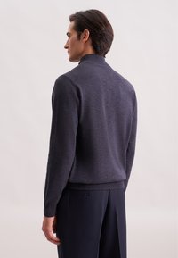 Uomo di lato che indossa un dolcevita grigio scuro e pantaloni scuri su uno sfondo chiaro e uniforme.