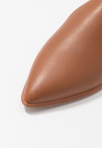 Pointe de chaussure en cuir marron à texture lisse avec couture visible le long du bord sur fond blanc.