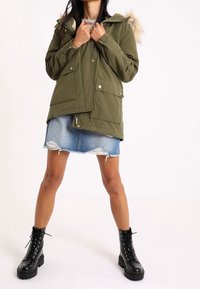 Olivově zelená parka bunda s kapucí lemovanou kožešinou, mnoha předními kapsami, zipovým zapínáním a volným střihem, ve spojení s džínovou sukní a černými botami.