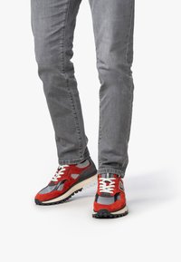 Scarpe da ginnastica rosse e grigie con materiali in mesh e camoscio, dotate di una suola testurizzata e lacci bianchi, abbinate a jeans slim-fit grigi.