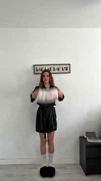 Crna saten haljina dugih rukava, zavezana u struku, uparena s bijelim čarapama i crnim pahuljastim papučama. Model drži laganu tkaninu.