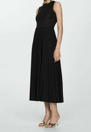 Kobieta w czarnej, plisowanej, bezrękawowej sukience midi i złotych, otwartych na palce wysokich butach na obcasie, stojąca na jasnym, jednolitym tle.