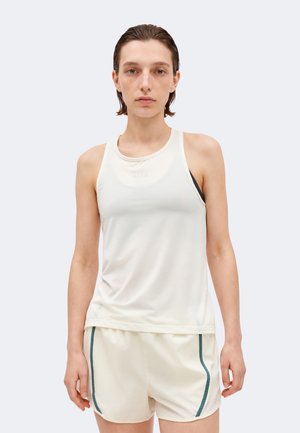 Άτομο με λευκό αμάνικο αθλητικό τοπ και ταιριαστά σορτς με σκούρες κάθετες ρίγες, που στέκεται μπροστά σε ένα μονόχρωμο φωτεινό φόντο.