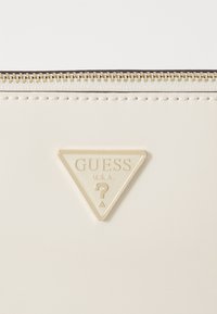 Portafoglio in materiale patent bianco con logo triangolare dorato e testo in rilievo "GUESS U.S.A." e un punto interrogativo. Dettaglio della cerniera dorata lungo la parte superiore.