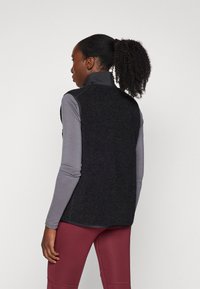 Black fleece vest with high collar, layered over a gray long-sleeve top. The model wears maroon fitted leggings, positioned from the back.