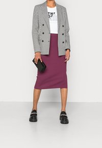 Vrouw in een zwart-wit houndstooth blazer, witte graphic t-shirt, paarse midi rok, zwarte instappers met kettingen, terwijl ze een zwarte clutch vasthoudt.