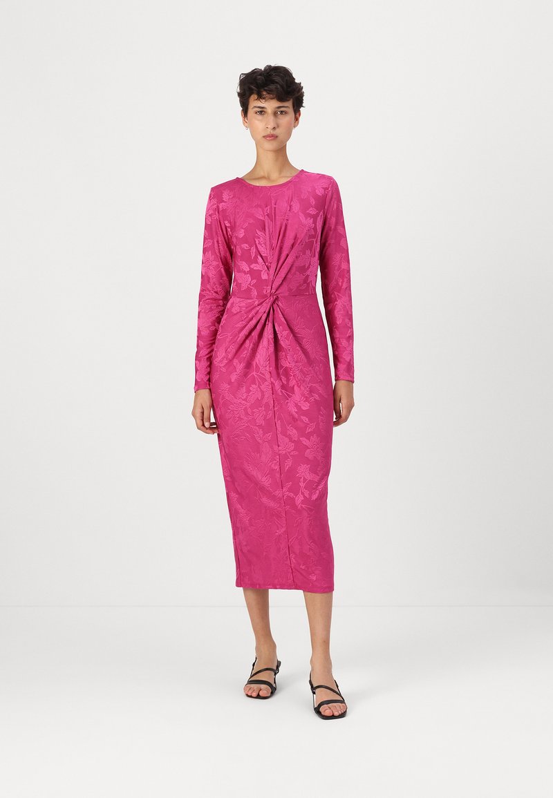 Vestido longo de mangas compridas em fúcsia com padrão floral texturizado, detalhe de cintura torcida e comprimento até à metade da panturrilha. Usado com sandálias pretas de tiras.
