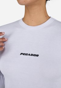 Hvit ribbestrikket langermet skjorte med rund hals og svart brodert tekst "PEGADOR" på brystet. Glatt tekstur, tettsittende design.