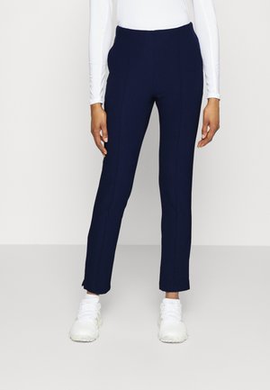 Personne portant un pantalon bleu marine ajusté, un haut blanc à manches longues et des baskets blanches, debout devant un fond clair uni.