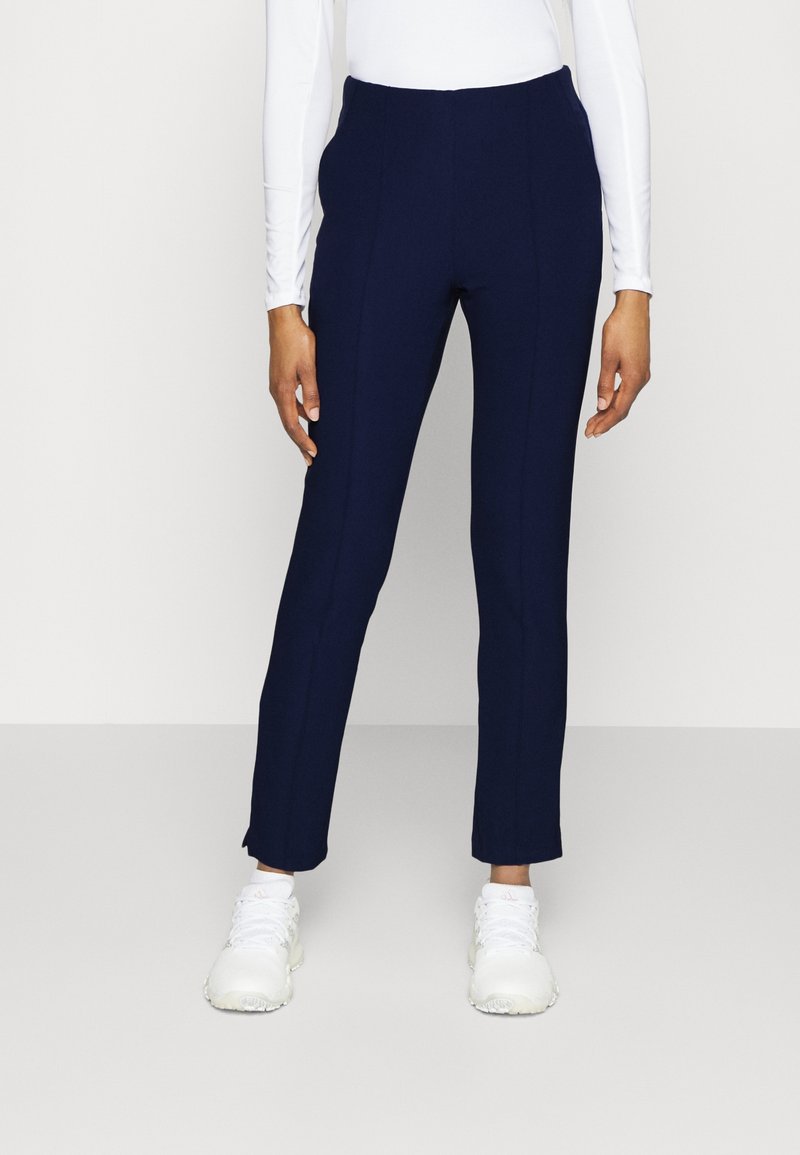Personne portant un pantalon bleu marine ajusté, un haut blanc à manches longues et des baskets blanches, debout devant un fond clair uni.