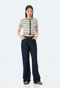 Cardigan corto a righe in crema e blu navy con scollo rotondo e chiusura a bottoni, abbinato a jeans a gamba larga blu scuro e scarpe marroni.