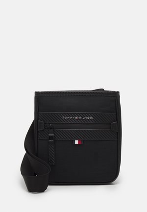 Schwarze Umhängetasche mit verstellbarem Riemen, vorderer Reißverschlusstasche und kleinem Tommy Hilfiger Logo in Rot, Weiß und Blau auf Textilmaterial.