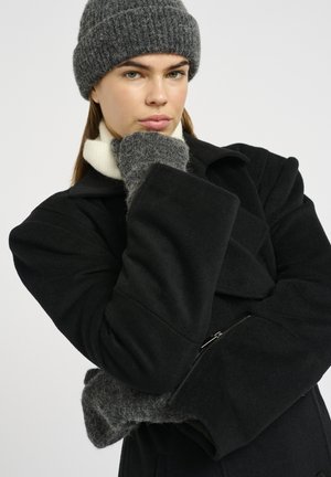 Black wool coat with large lapels and zipper pocket, paired with a grey textured knit hat and matching gloves. White mock neck sweater underneath.
