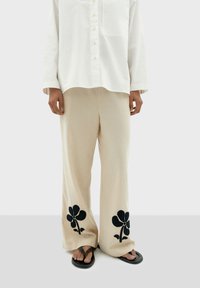 Pantaloni beige a gamba larga con ricami di fiori neri sui bordi inferiori, abbinati a una camicia bianca morbida e infradito scuri.