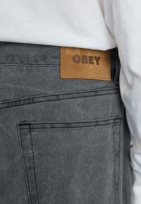 Šedé denimové nohavice s koženou nášivkou označenou "OBEY" na zadnej strane, s klasickým prešívaním vreciek a hladkým povrchom.