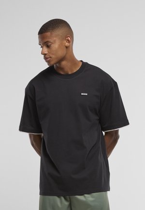 Nuori mies, jolla on lyhyet hiukset ja joka käyttää mustaa ylisuurta t-paitaa, jossa on pieni "KANI"-logo rinnassa, katsoo vasemmalle.