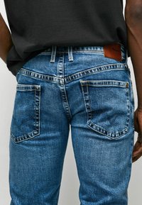 Vaqueros de mezclilla azul con un corte clásico, cinco bolsillos, detalles de costura y una etiqueta de cuero con la marca en la cintura.