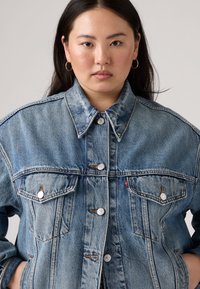 Light blue denim jacket with a collared neck, buttoned front, two chest pockets, and contrasting stitching. Soft texture, loose fit.