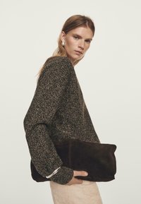 Kobieta w ciemnym swetrze z dzianiny i kremowych spodniach, trzymająca dużą ciemno-brązową torebkę, spoglądająca przez ramię na jednolitym tle.