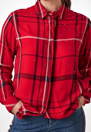 Chemise à manches longues rouge à carreaux avec des lignes blanches et foncées, petits clous argentés, portée rentrée dans un jean bleu avec un détail rayé sur le côté.