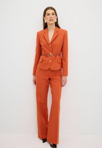 Op maat gemaakte oranje blazer met gouden knopen en zijzakken, gecombineerd met bijpassende broek met wijde benen. Bruine riem op de taille. Soepele textuur.