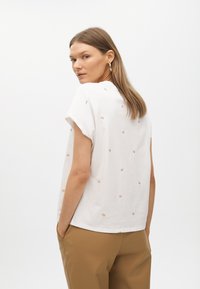 Haut blanc à manches courtes avec des broderies "W" légères de couleur brun clair, présentant un col rond et une coupe décontractée. Associé à un pantalon beige.