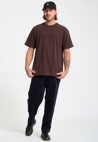 T-shirt en coton marron à manches courtes et col rond. Associé à un pantalon en velours côtelé bleu marine et des chaussures noires à lacets. Casquette noire sur la tête.