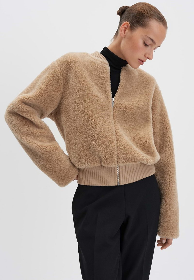 Giacca croppata beige e fluffy con zip frontale, bordo a coste e maniche lunghe. Indossata sopra un collo alto nero, che mette in evidenza una superficie texturizzata.