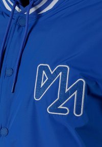 Mėlynas nailono švarkas su baltu juostotu apykakle, su ryškiu baltu siuvinėtiniu logotipu ir apvaliu mygtuku priekyje.