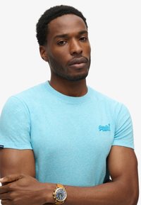 T-shirt azzurro a maniche corte con una sottile texture e logo ricamato sul petto. Indossato con un orologio dorato e le braccia incrociate.