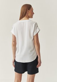 Camisa blanca de manga corta con puños doblados, dobladillo ligeramente curvado y aberturas laterales, presentada con pantalones cortos azul marino, textura minimalista y suave.