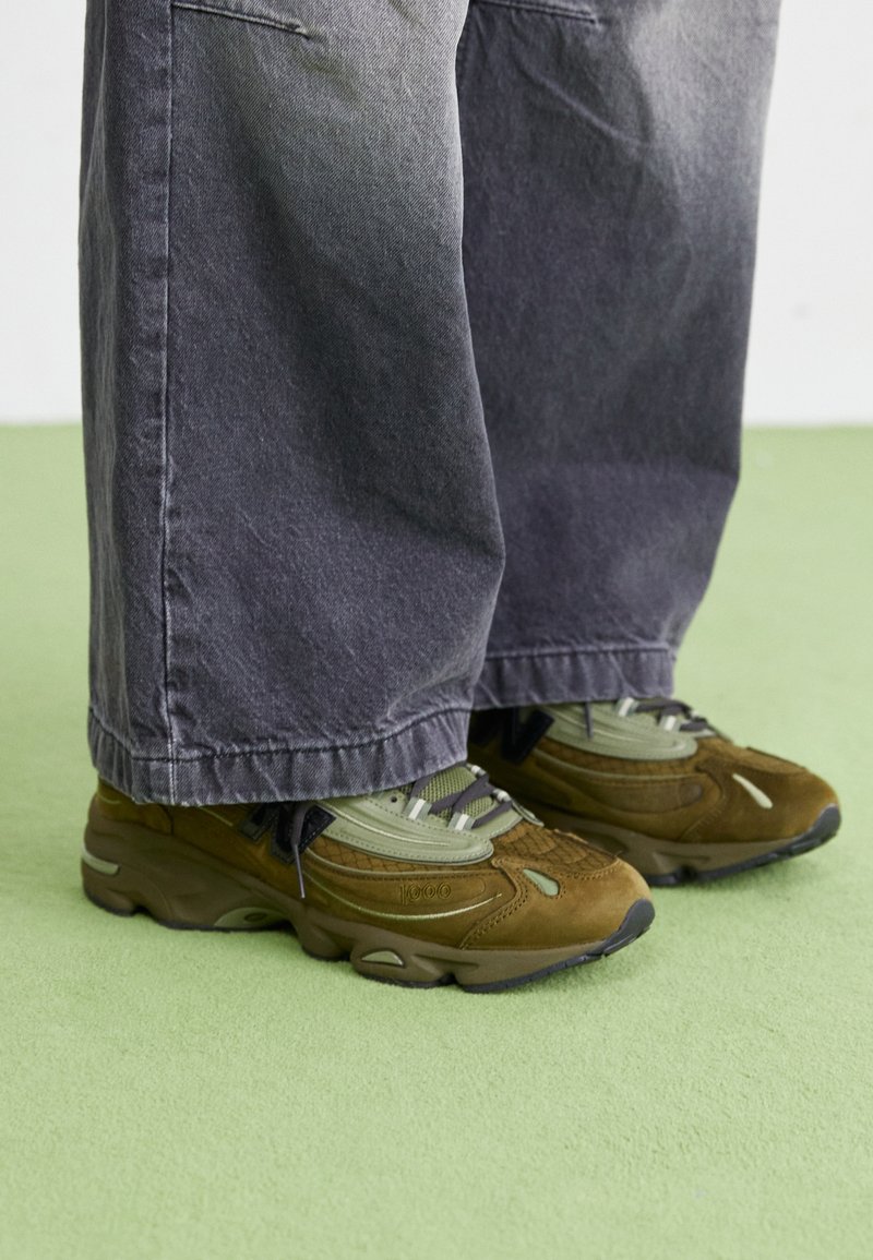 Personne portant un pantalon gris large et des baskets marron et vertes debout sur un sol vert clair.