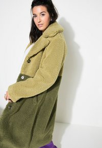 Cappotto lungo oversize bicolore realizzato in un morbido materiale testurizzato. Parte superiore di colore verde oliva, parte inferiore di una tonalità più chiara. Chiusura frontale con bottoni.