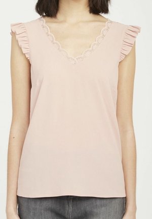 Kvinde iført en ærmeløs lyserød top med V-hals med blonder og rynkede skulderdetaljer, kombineret med mørkegrå jeans.