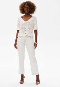Biała dzianinowa bluzka z dekoltem w serek i krótkimi rękawami, ozdobiona wzorem liści i drewnianymi guzikami. Połączona z jasnymi jeansami i szpilkami.