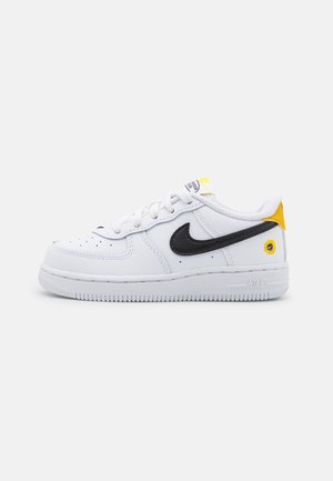 Baskets basses blanches Nike avec un logo Swoosh noir, des accents jaunes sur le talon et la languette, et une zone d'orteils perforée.