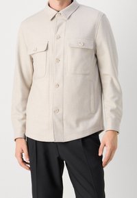 Chaqueta beige de botones con dos bolsillos en el pecho y bolsillos laterales, hecha de tejido suave. Presenta un collar clásico y un dobladillo recto.