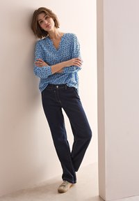 Blusa blu a fantasia con scollatura a V, abbinata a jeans in denim scuro. La modella indossa scarpe chiare, appoggiata a un muro.