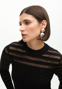 Čierny dlhý rukáv top s priesvitnými horizontálnymi pruhmi, vysokým golierom a malým srdiečkovým logom. Veľké kovové náušnice v zlatých a strieborných tónoch.